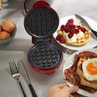 Red waffle maker open on tabletop, with plates of waffles, raspberries, cream, bacon and fried egg