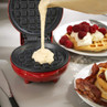 Red waffle maker with batter being poured in, plates of waffles and bacon beside