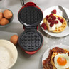 Red waffle maker open, surrounded by eggs, flour, and plated waffles with raspberries, cream, and bacon.