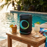 Black portable speaker with LED ring, shown on table by pool with tropical drinks