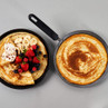 Two frying pans with large pancakes, one topped with mixed berries, banana and cream.