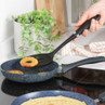 Black slotted spatula lifting a cooked pineapple ring from non-stick frying pan on hob