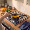 Hand stirring soup in black saucepan on gas hob, butternut squash and herbs on worktop