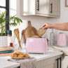 Pink two-slice toaster on kitchen counter, hand adding seeded bread, matching kettle in background
