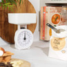White mechanical kitchen scale on worktop beside flour, eggs and cookbooks