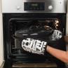 Person placing large black roasting pan in oven with black and white utensil oven gloves