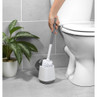 Grey toilet brush with holder beside white toilet on tiled bathroom floor