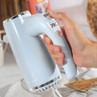 Hand holding pale blue electric hand mixer with speed controls, shown in kitchen setting