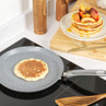 Grey non-stick pancake pan on hob with pancake, stack of pancakes and peaches beside