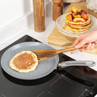 Grey frying pan on induction hob with pancake, stack of pancakes and fruit in background