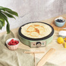 Electric crepe maker in green cooking a pancake, shown with fresh fruit and baking ingredients.