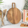 Round acacia wood serving board on kitchen counter with utensils and salt and pepper pots