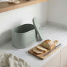 Light green ribbed metal bread bin with lid, shown next to sliced loaf on board.