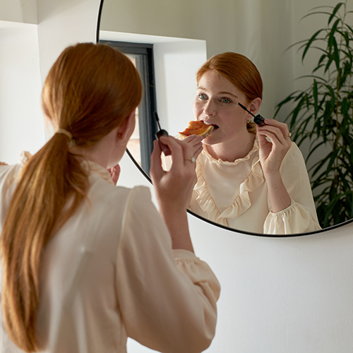 Woman eating toast and putting on makeup in mirror