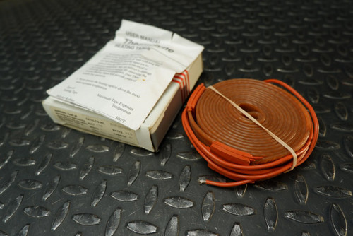 An unused BriskHeat BS0102080 silicone rubber heating tape, 1 inch by 8 feet, 240V, 418W, is displayed coiled in a vibrant reddishorange color and secured with a light beige rubber band. Beside the coiled heating tape sits its partially opened white cardboard product box, revealing the "USER MANUAL" and "HEATING TAPE" text, all resting on a dark, reflective diamond plate metal surface. This new BriskHeat heating tape is clearly visible and ready for heat tracing applications.