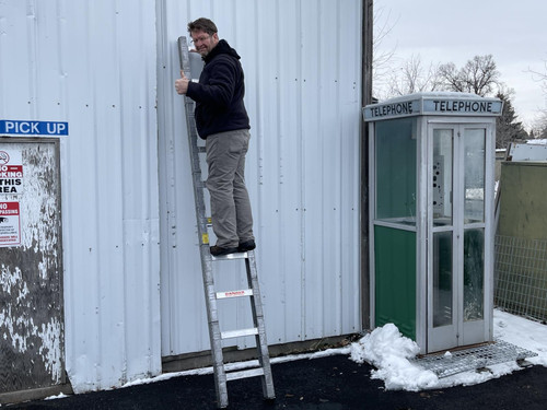 A man in casual attire gives a thumbs-up while standing on a tall, quickstep folding tactical ladder leaned against a white metal container. Beside him, there is a standalone, vintage telephone booth on snow-dusted ground. The setting conveys a chilly, utilitarian outdoor area with industrial vibes.