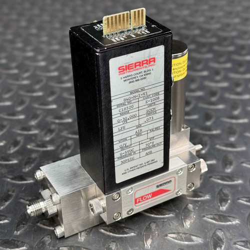 Sierra Mass Flow Controller 840M1V1, capable of measuring 030,000 SCCM for Air, prominently displayed on a dark textured surface. The industrial instrument features a black rectangular top housing with a white product label detailing its model number, serial C18520, range, and gas type, along with the Sierra Instruments logo and company address. Gold electrical connector pins are visible on the top of the black enclosure. Below, a silvertoned, brushed metal manifold block is visible, featuring a "FLOW" indicator with a red arrow and various stainless steel fittings and screws for gas connections, highlighting its robust design for precise gas flow control applications.