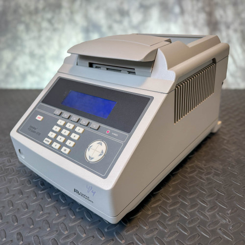 Applied Biosystems GeneAmp PCR System 9700, 96-Well Thermocycler from Obtainium Science and Surplus shown at an angle, displaying the keypad, screen, and lid on a textured metal surface.
