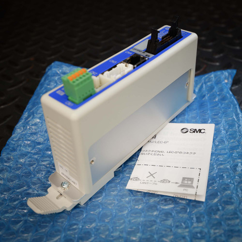 Unused SMC LECP6 Series Step Motor Controller, model LECP6PDLEHF20K248, a white and blue industrial automation module. The controller features a green screw terminal connector, multiple RJ45 Ethernet ports (CN3, CN4), and a black multipin connector on its top edge. A metal mounting bracket with a thumbrelease lever is visible on the side. The device rests on blue bubble wrap, with an SMC instruction manual detailing LAN cable connection diagrams next to it, emphasizing its new condition for motion control applications.