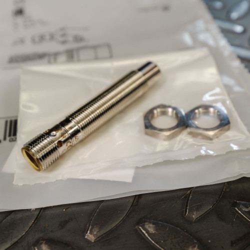 IFM sensor body with intact threads, no visible wear or damage. Two mounting nuts in clear plastic bag.