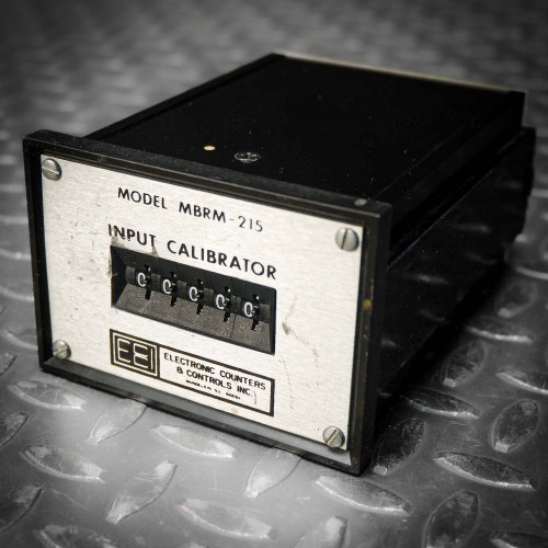 "A black ECCI MBRM-215 input calibrator module on a textured gray surface, featuring a front label with white text specifying model details and serial number '865343.'