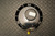 Rear view of an Ashcroft pressure gauge featuring a black, ribbed 6-inch diameter housing and a central circular cover plate, showing the bottom brass inlet fitting capped with a yellow protective plug.