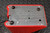 Red housing with perforated metal top panel, four dome-head screws visible. Black cable exits lower-left edge. Minor surface wear on housing corners.