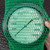 A round gauge or dial is being held inside a green Poly-Net HDPE Protective Mesh Netting Tube from Obtainium Science and Surplus.