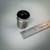 Cylindrical stainless steel body with satin finish; top face marked 50 g on polished black surface. Ruler shows approximately 1-inch diameter.