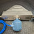 A close-up of the back view of the Micro Motion Coriolis flow meter from Obtainium Science and Surplus, showing the blue sensor attached at the center. The device rests on a metal floor with a diamond-pattern texture, and a blue cable extends from the sensor.