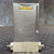 Matheson 82720412 Mass Flow Transducer, 100 SCCM, Air. This precision industrial flow meter features a durable, textured silvergray metal casing. At the top, a recessed gold electrical connector with labeled pins, including 'L' and 'A', is visible for data and power. The bottom section of the sensor shows a brushed silver metal block equipped with two threaded fluid fittings on either side, designed for secure air connections and accurate flow measurement in industrial equipment.