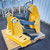 Yellow-painted steel frame with visible bolt hardware, pivot pins, slotted mounting feet, hydraulic cylinder, and black hydraulic hoses with fittings attached.