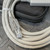 A closeup shot showcases the SMC LECW2 Communication Cable, a light gray industrial data cable with a clear RJ45 connector and a durable gray strain relief boot. The cable is neatly coiled and secured with a white strap, displaying visible specifications such as "AWM" and "LL65413". It rests on a dark gray diamond plate patterned surface, with a black textured zippered carrying case and a corner of a gray USB converter unit (for the LATCA Controller) visible in the background, emphasizing the complete connection solution.