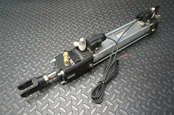SMC CDLADN50250EK39XC18 pneumatic actuator air cylinder, shown horizontally on a dark grey diamond plate background. The robust silvergrey rectangular aluminum body is visible, flanked by black end caps. A black clevis mount rod end is prominently featured at the front, attached to the metallic piston rod. Two black sensor switches with bundled electrical wiring are mounted along the side of the cylinder body. Several brass and white pushtoconnect pneumatic fittings are visible on the black control blocks, indicating air input and output ports. This industrial automation component is a doubleacting air cylinder.