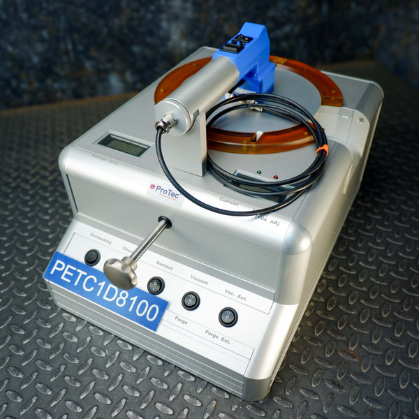 Close-up of a ProTec electrostatic wafer handling chuck racket on a metal surface, showcasing its control panel with buttons for de-chucking, chucking, contact, vacuum, vacuum exhaust, and purge, with a blue and silver handheld wafer handling racket resting on top.