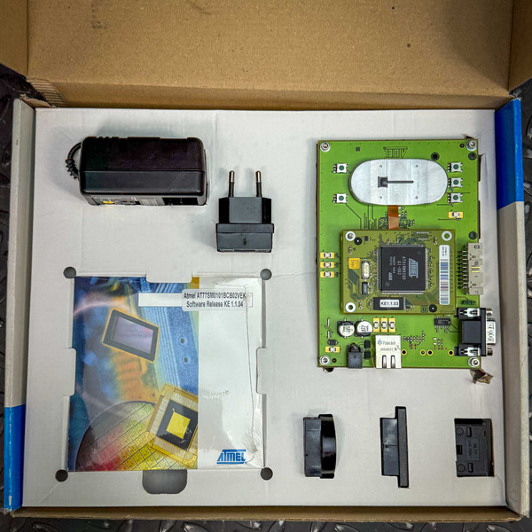 A top-down view of the ATMEL Biometric FingerChip development board kit from the Obtainium Science Surplus Store, showing various components neatly arranged in a box, including a green circuit board, a power adapter, and connectors.