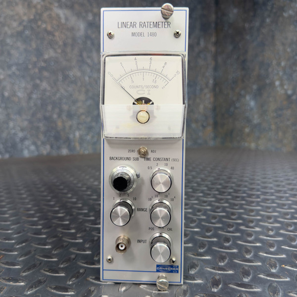 Front view of a Canberra 1480 Linear Ratemeter for Radiation Detection and Nuclear Physics from Obtainium Science and Surplus. The device has a white face with a needle gauge measuring counts per second. Several dials and inputs are below the gauge for adjusting settings like range and time constant. The background is a textured metallic surface.
