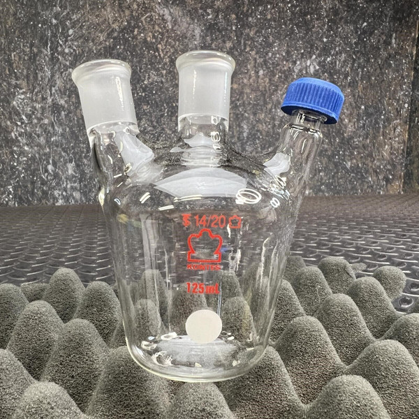 A clear glass European-Style 3-neck flask from Obtainium Science and Surplus, with two glass stopper necks and one threaded blue cap. The flask rests on a foam-like surface with a textured dark background.