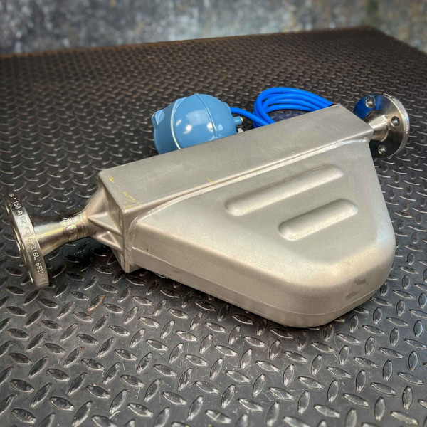 A metallic Micro Motion Coriolis flow meter from Obtainium Science and Surplus with a triangular shape, featuring a smooth, ridged surface, and a cylindrical connection on one end. A blue sensor and cord are attached at the top, resting on a diamond-patterned metal floor.