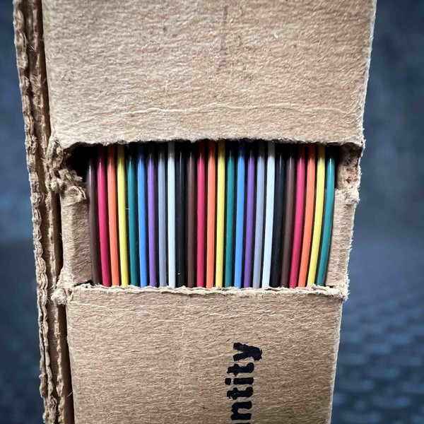 Detailed view of a Belden Flat Ribbon Cable 9R28025 28AWG 25C Unshielded, showcasing 25 vibrant, multicolored insulated wires neatly arranged in a flat array, visible within a rectangular opening of its brown corrugated cardboard packaging. The diverse wire colors include shades of brown, red, orange, yellow, green, blue, purple, gray, white, and black. This closeup highlights the organized presentation of the highquality data cable, commonly available in 100ft spools, ideal for electronic projects requiring organized wiring.