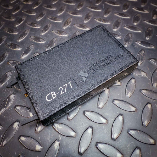 Angled top view of the National Instruments CB-27T Isothermal Terminal Block from Obtainium Science and Surplus. The black textured casing features an embossed "National Instruments" logo and a label with "CB-27T" in bold white text. The device is resting on a diamond-plate metal surface.