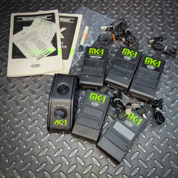 Surplus lot of MK-1 Audio Dosimeters and AC-1 Acoustical Calibrator from Obtainium Science and Surplus displayed on a textured metal surface. The set includes four black MK-1 audio dosimeters labeled with green text, an AC-1 acoustical calibrator with a dial for decibel adjustment, multiple microphone attachments with cables, and operating manuals from DuPont.