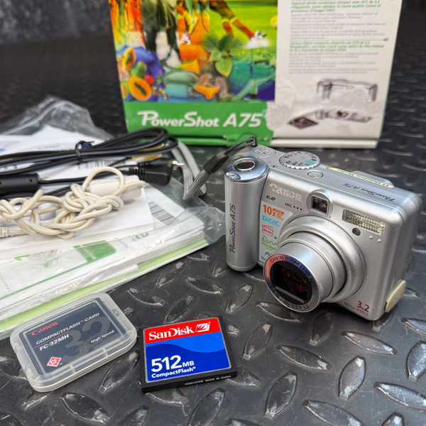 Vintage Canon PowerShot A75 3.2MP Digital Camera from Obtainium Science and Surplus shown with original packaging, two CompactFlash memory cards (one 32MB Canon and one 512MB SanDisk), connection cables, user manuals, and camera accessories displayed on a textured black surface.
