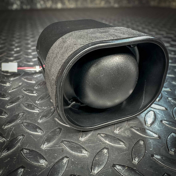 Close-up of an RSG Emergency Vehicle Siren from PartsMine.com, showing the front view with a black protective grill and a cylindrical body, placed on a textured black metal surface.