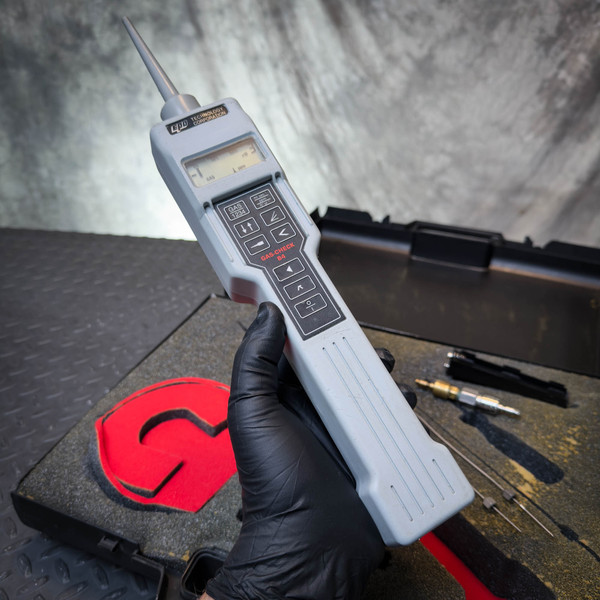 EPD Technology Digital Gas Leak Detector, B4 Gas Check shown held in a gloved hand above its foam-lined carrying case, featuring a long gray body with display and keypad.