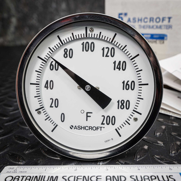 Closeup of an Ashcroft 50EI42E060 bimetal thermometer, showcasing its 5inch white dial with a 0200°F temperature range and clear black numerals. The black indicator needle points precisely to 70°F. The thermometer features a reflective stainless steel bezel and prominently displays the Ashcroft brand logo and '°F' symbol. This industrial or laboratorygrade temperature gauge is set against a dark, textured background, with a ruler from Obtainium Science and Surplus and product packaging partially visible, indicating a reliable temperature measurement instrument.