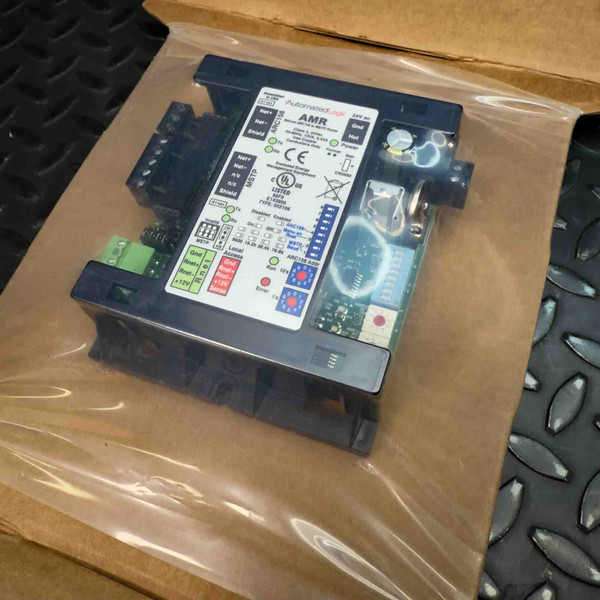 Closeup of an unused Automated Logic AMR BACnet ARC156 to MS/TP Router, ARCNET BT485 communication module, still sealed in its clear plastic packaging within a brown cardboard box. The black rectangular device features a prominent label detailing "Automated Logic AMR ARC156", input/output terminals for network connections including Net, Net, Shield, Gnd, Rn A, Rn B, and 12V. Visible green and black connectors, status indicator LEDs (green, red, yellow), and certification markings like CE and UL LISTED are clearly displayed on this building automation network interface for HVAC control systems, appearing pristine and ready for installation.