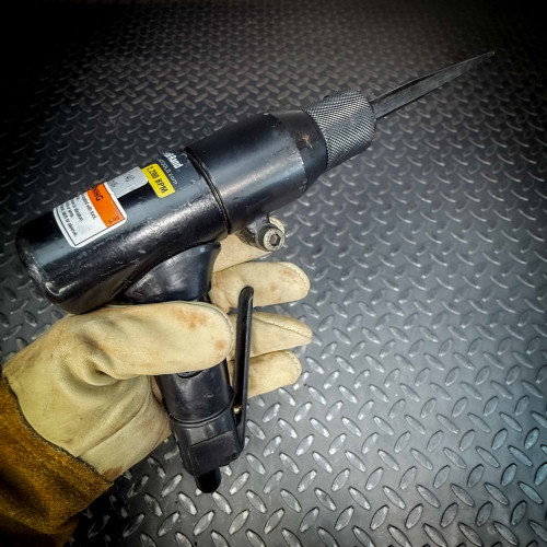 A close-up of an Ingersoll Rand 180PG Pistol Grip Chisel Scaler held in a gloved hand, placed on a diamond-plate metal surface. The tool features a chisel attachment and warning label.