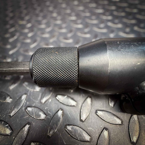 A side view of the Ingersoll Rand 180PG Pistol Grip Chisel Scaler, focusing on the textured grip and chisel mechanism against a metallic diamond-plate surface.
