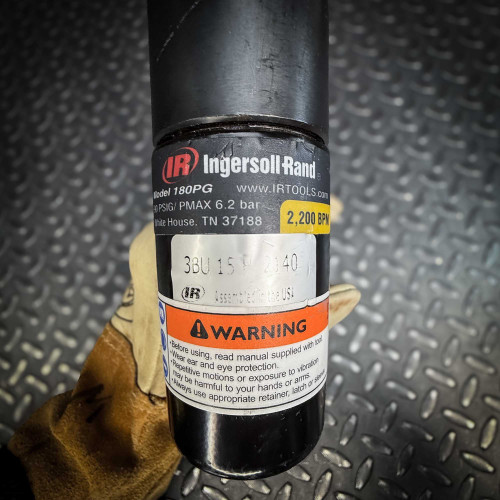 A detailed view of the Ingersoll Rand 180PG Pistol Grip Chisel Scaler label, displaying the brand logo, specifications, and safety warning. Held in a gloved hand on a diamond-plate surface.